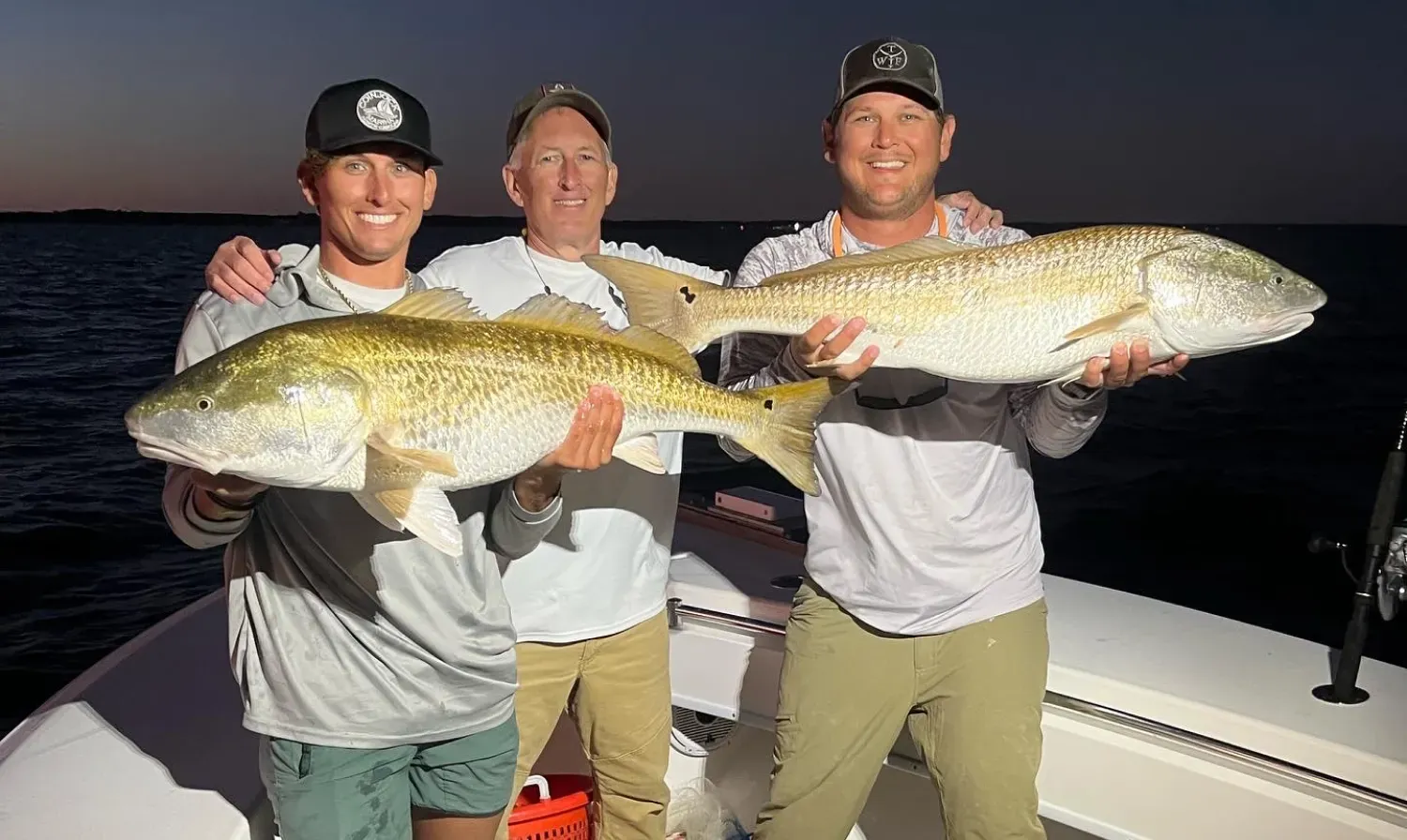 exp-1 Three smiling men on a boat hold two large fish at dusk, with water and a dark sky in the background.
