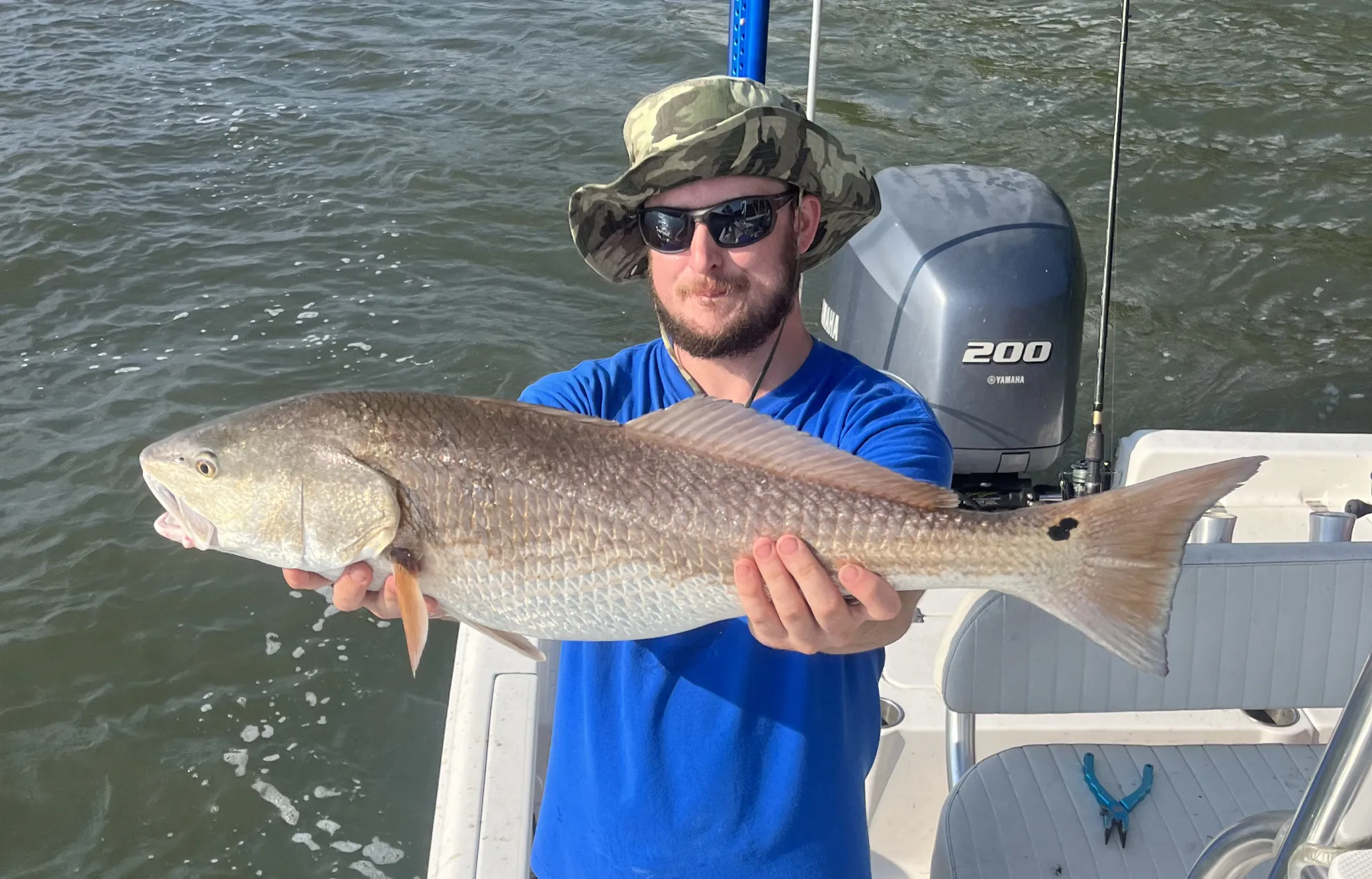 red-drum-1 Man in a blue shirt and hat holds a large fish on a boat, with water in the background.