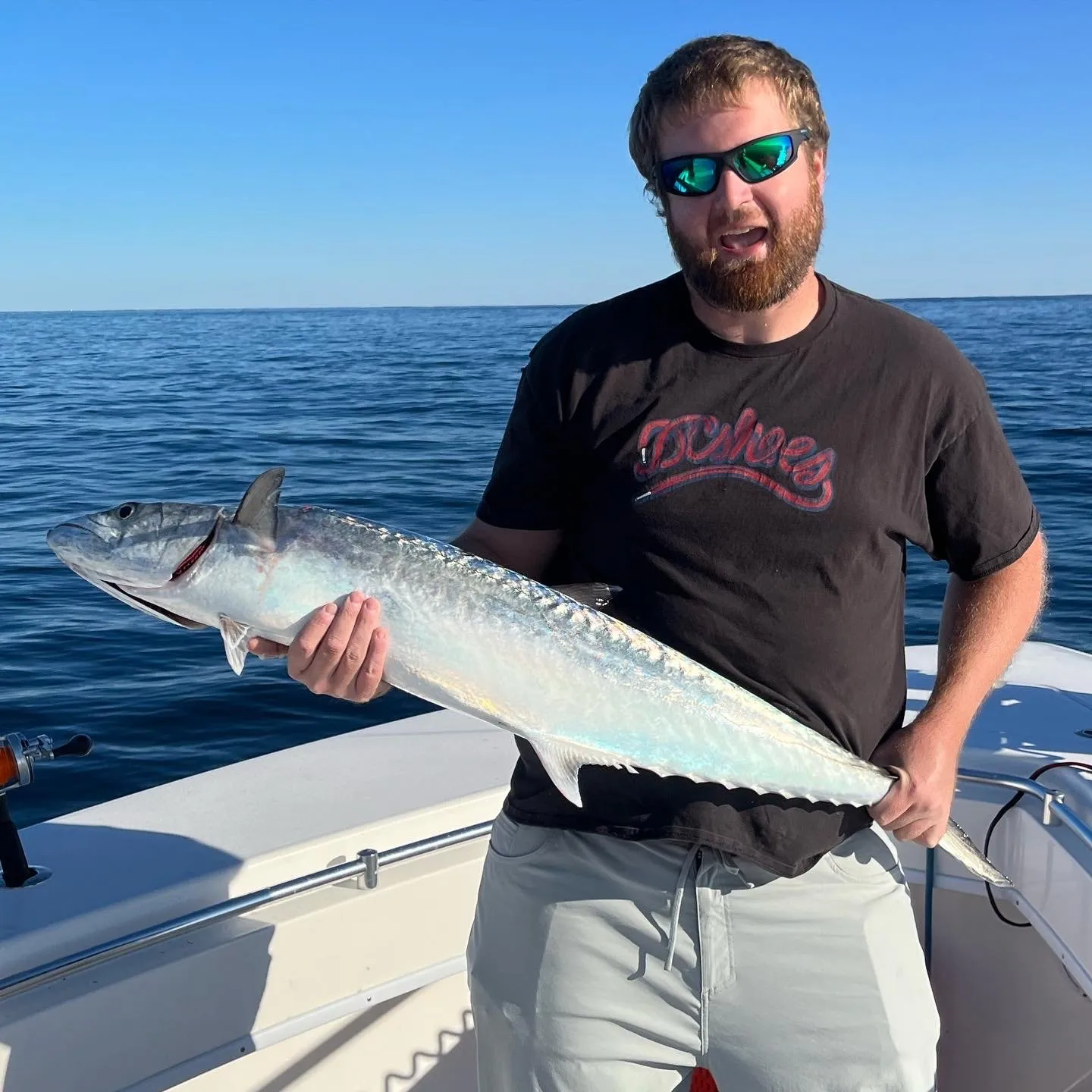 king mackerel-1 Man on a boat holding a large fish, wearing sunglasses and a black t-shirt, with the ocean in the background.