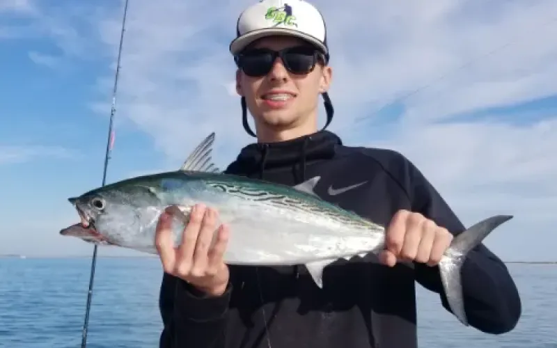 Young man in sunglasses and a hat holding a freshly caught fish on a boat with water in the background.