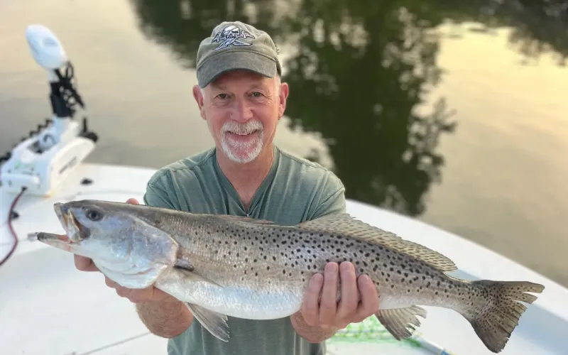 Smiling man on a boat holding a large speckled fish with both hands near the water.