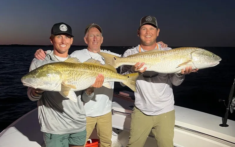 Three men on a boat at dusk proudly holding large fish they caught, smiling at the camera.