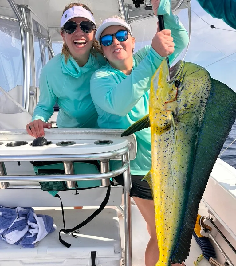 Two women on a boat smiling and holding a large, colorful fish they caught while fishing.