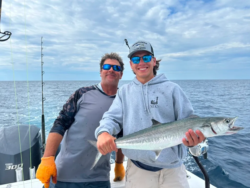 about-h Two people on a boat holding a large fish, smiling, with ocean and cloudy sky in the background.