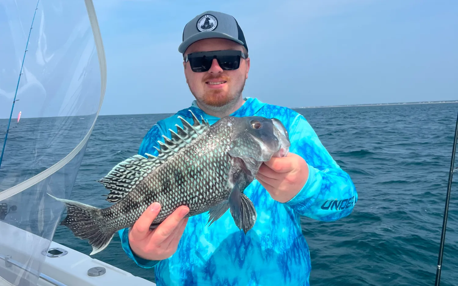 Man in blue shirt and hat holding a large fish on a boat with the ocean in the background.