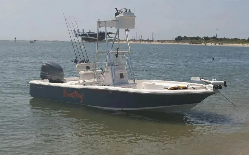 A fishing boat with multiple rods is anchored near the shore in shallow, calm water under a clear sky.