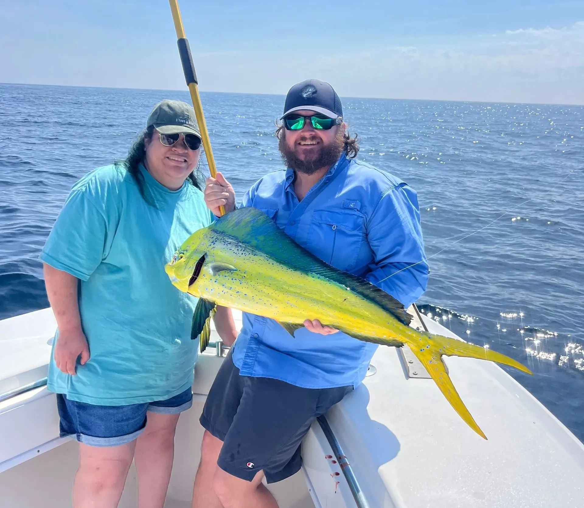 Two people on a boat smiling while holding a large, bright yellow and green fish with the ocean behind them.