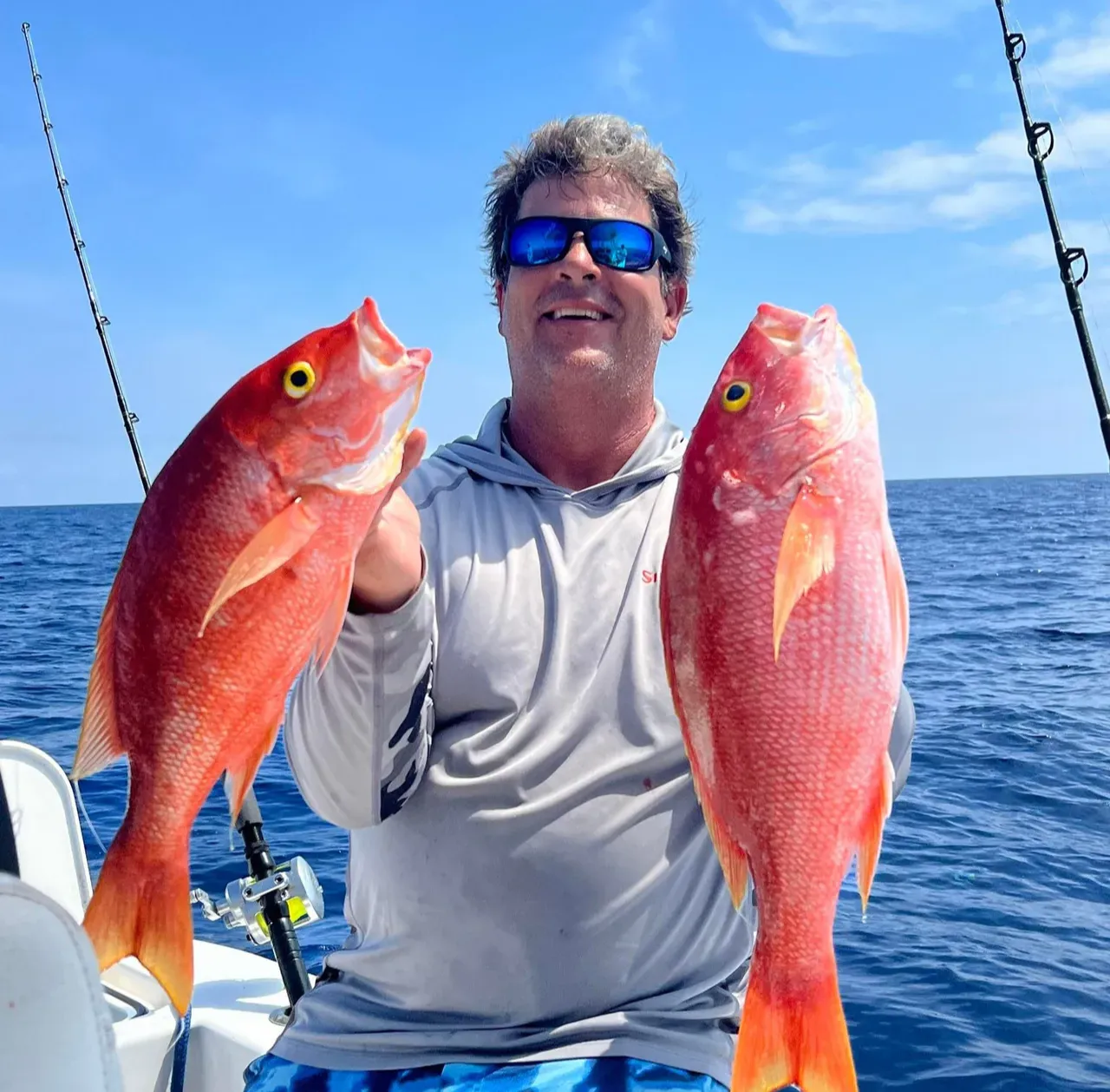 Man on a boat holding two large red fish, with the ocean and blue sky in the background.