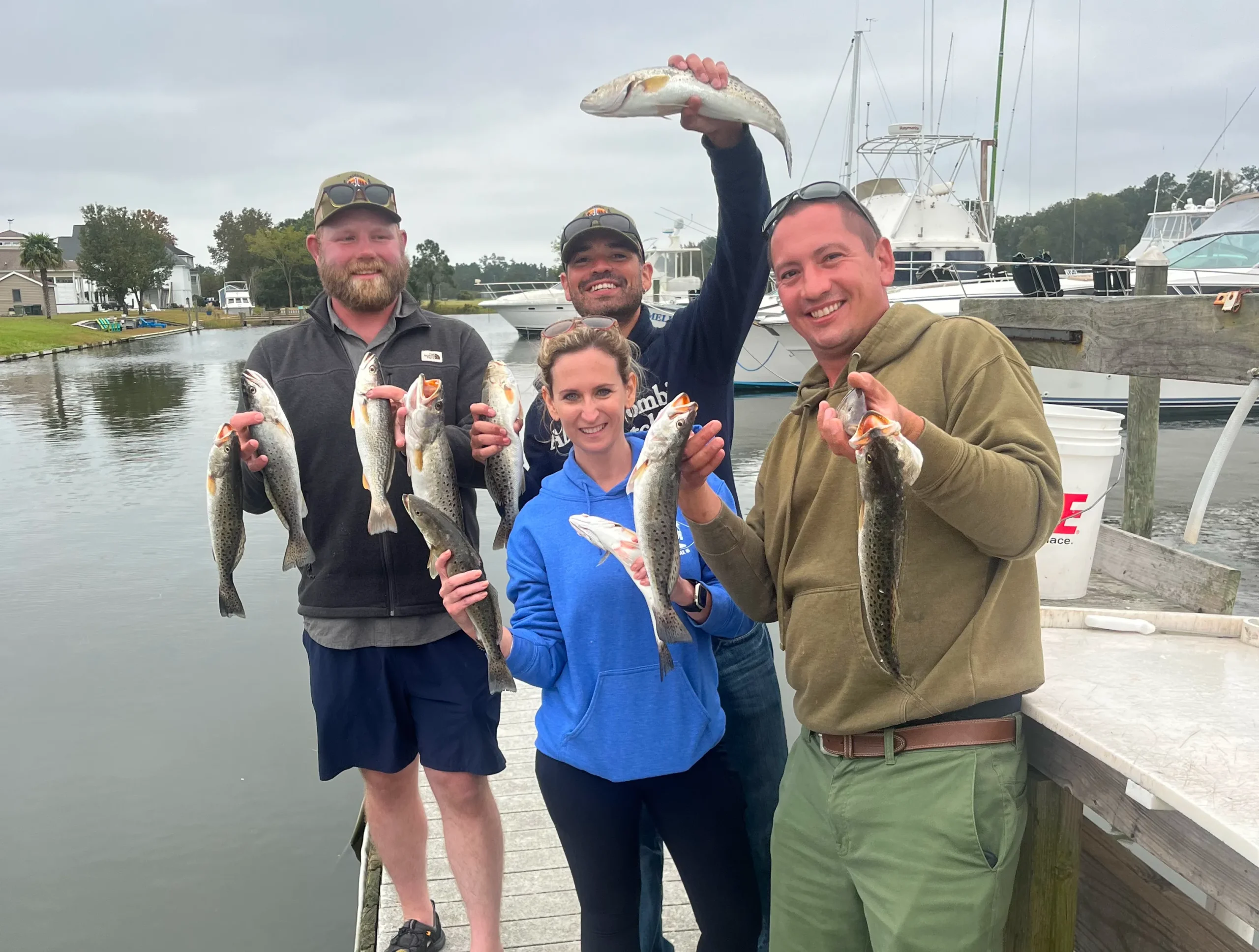 charters-1 Four people on a dock proudly holding several fish they caught, with boats and water in the background.
