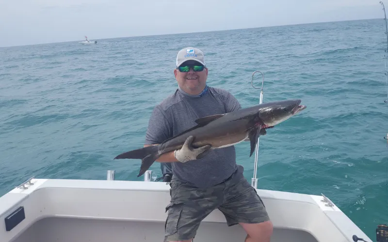 Man on a boat holding a large fish, smiling at the camera with the ocean in the background.