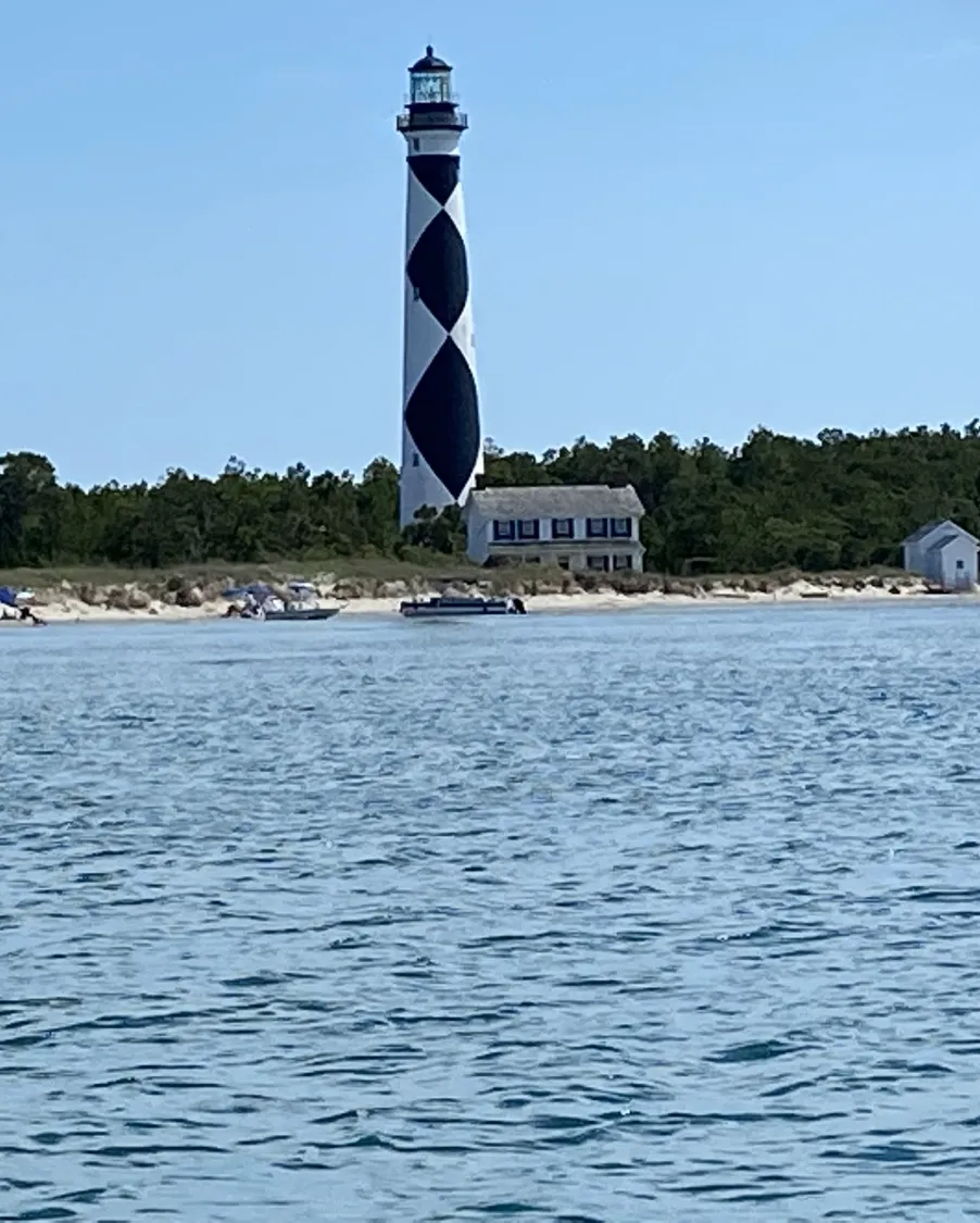 A tall lighthouse with a black-and-white diamond pattern stands by a beach, with water in the foreground.