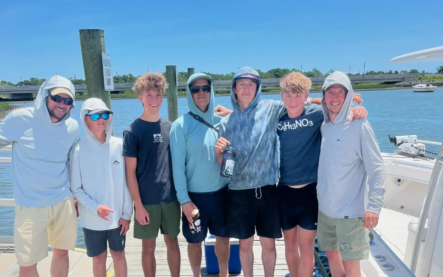Seven people in casual summer clothes stand on a dock by the water, smiling on a sunny day.