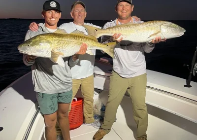 Three men on a boat at dusk smiling and holding two large fish they caught.