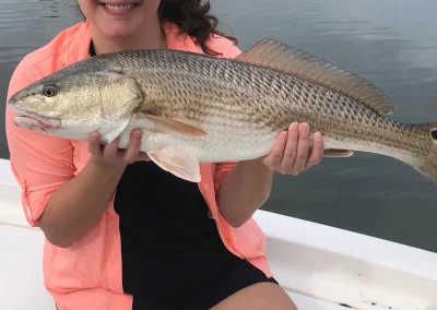 Smiling woman on a boat holding a large fish, wearing a pink shirt, black shorts, and a visor.
