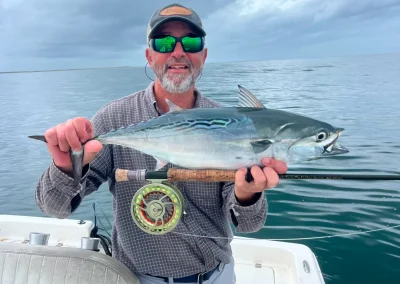 Man on a boat holds a large fish and a fishing rod, smiling with sunglasses under a cloudy sky.