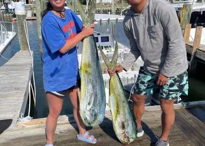 Two people on a dock hold large fish, with boats and a marina in the background. Another fish lies on the dock.
