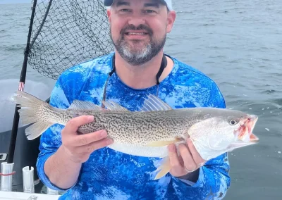 Man in a blue shirt holding a large fish on a boat, with water and a fishing net in the background.