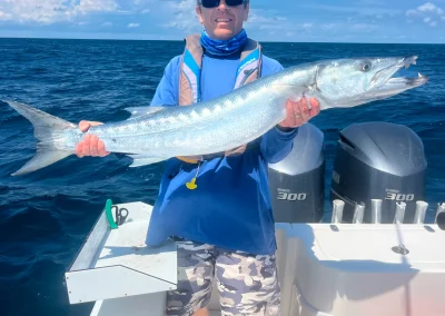 Person on a boat holding a large barracuda fish over blue ocean water, with engines visible in the background.