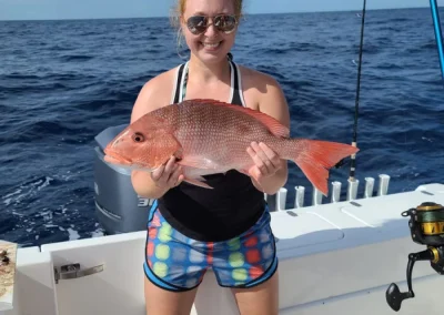 Smiling woman on a boat holding a large red fish with the ocean and fishing rods in the background.