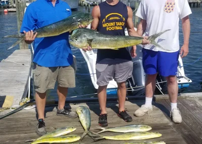 Three men on a dock holding large fish, with several more fish laid out in front of them. Boats in background.