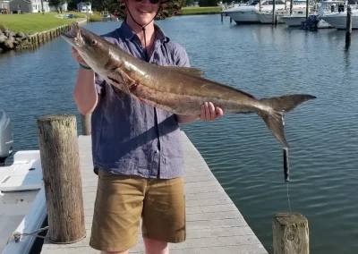 Smiling person in a hat holding a large fish on a dock with boats and water in the background.