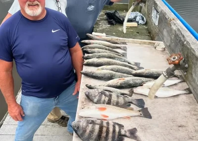 Three smiling men on a dock display freshly caught fish lined up on a table near the water.