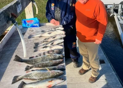 Two men on a dock smile beside a row of caught fish laid out on a table next to a truck.