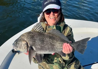 Smiling woman in camo holds a large fish on a boat with water in the background.