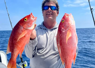Man on a boat holding two large red fish, smiling, with fishing rods and blue ocean in the background.