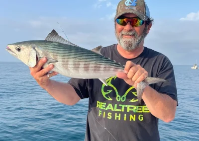Man in sunglasses and a cap holding a striped fish on a boat with the ocean and sky in the background.