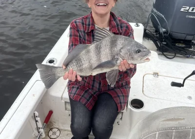 Smiling person in a plaid shirt holding a large fish on a boat by the water.