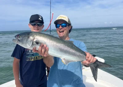 Two smiling men on a boat hold a large fish, with ocean water and blue sky in the background.