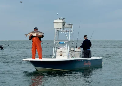 Man in orange overalls holds a large fish on a boat, another man fishes nearby in open water under a cloudy sky.