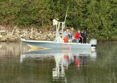 A small boat with four people on board floats near a wooded shoreline on calm water.