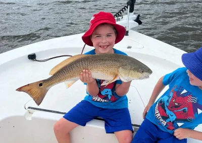Smiling child in a red hat holds a large fish on a boat next to another child wearing a blue hat.