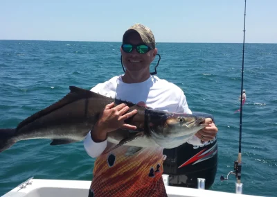 Smiling person on a boat holding a large fish with the ocean and fishing rod in the background.