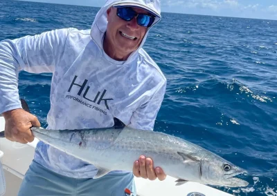 Man on a boat holding a large fish, smiling, wearing a hoodie and sunglasses, with ocean in the background.