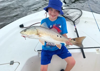 Smiling child in a blue hat proudly holds a large fish while sitting on a boat with fishing gear nearby.