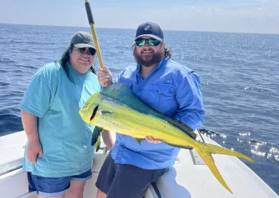 Two people on a boat holding a large yellow and green fish with the ocean and sky in the background.