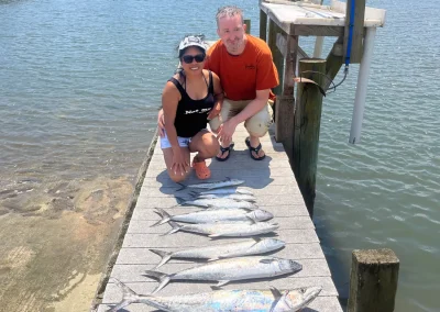 Two people kneel on a dock beside seven large fish they caught, with water and boats in the background.