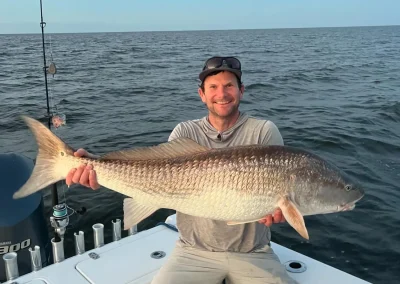 Man smiling on a boat, holding a large fish with both hands over his lap, ocean in the background.