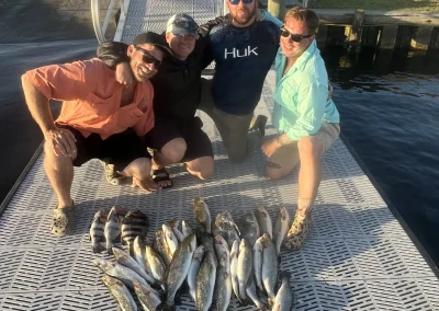 Four people smiling on a dock with a large catch of fish laid out in front of them on a sunny day.