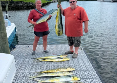 Two people on a dock proudly holding large fish, with several more fish laid out in front of them.