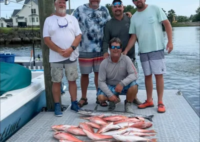 Five men on a dock posing with a large catch of fish laid out in front of them, boat and houses in background.