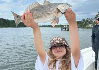 Smiling child on a boat holding up a large fish with both hands, water and trees in the background.