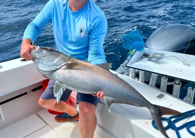 Man smiling on a boat, holding a large fish with the ocean and blue sky in the background.