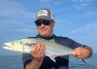 Man wearing sunglasses and a cap holding a large fish on a boat under a blue sky.