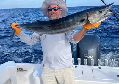 Man in sunglasses and hat holding a large fish on a boat with fishing rods, ocean in the background.