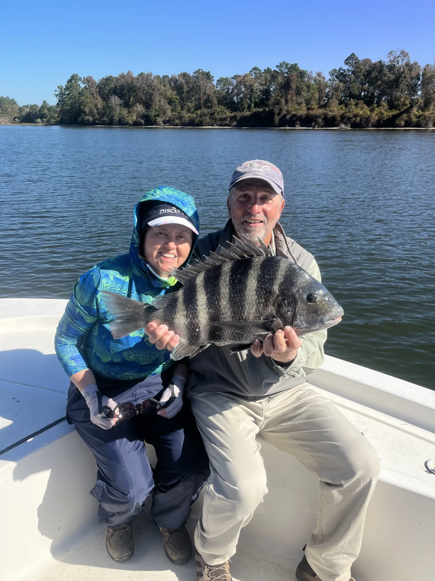 Two people on a boat smiling and holding a large black-and-white striped fish by a lake.