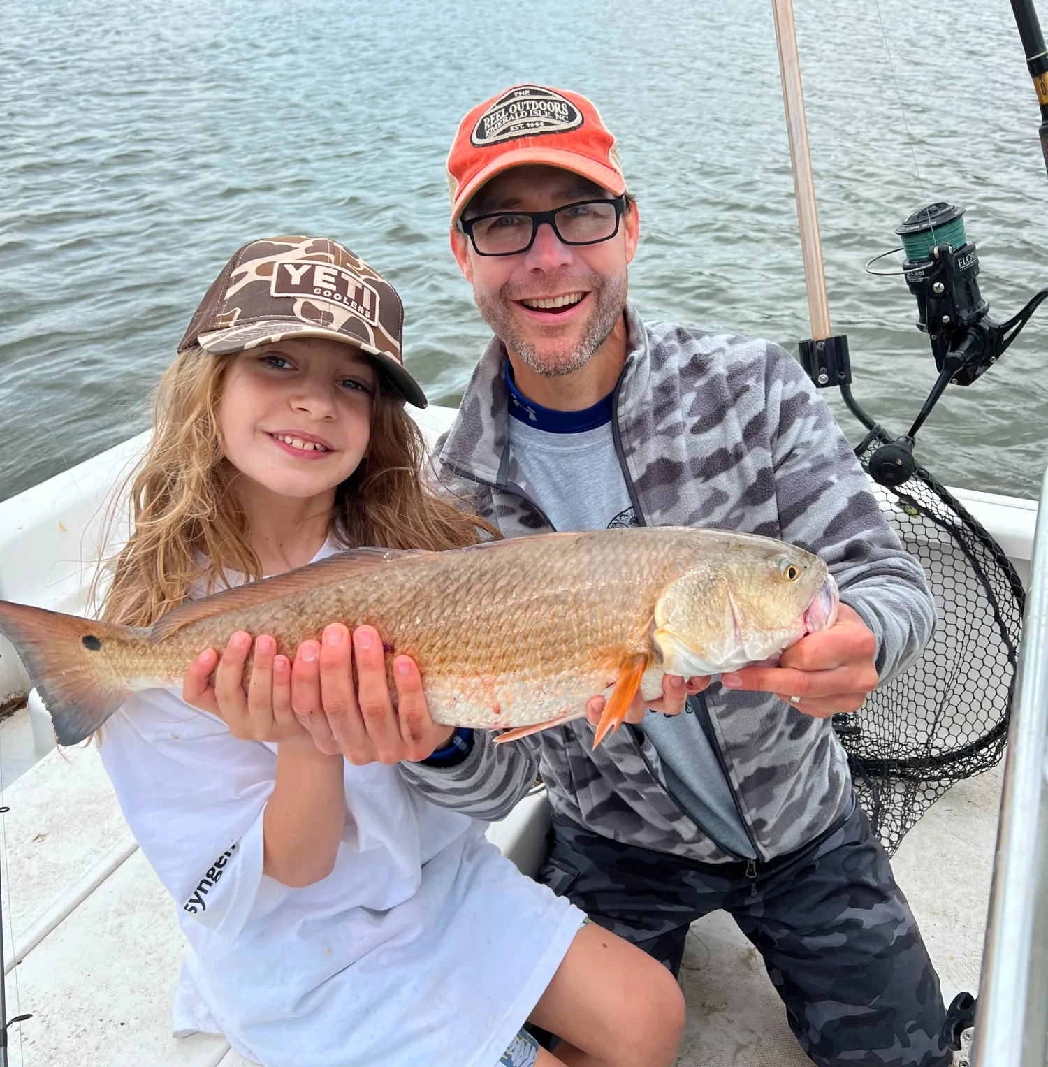 Smiling man and child on a boat holding a large fish they caught, with water in the background.